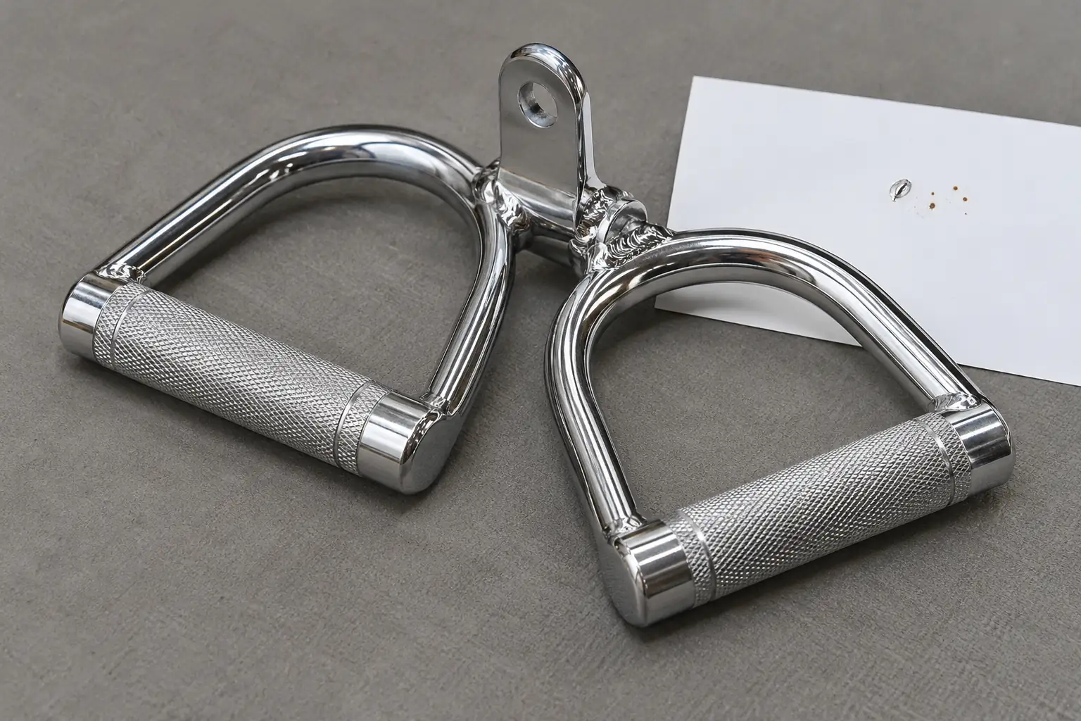 Accesorio cromado de doble agarre sin marca en una mesa de inspección con tarjeta blanca y pequeños indicios de corrosión superficial